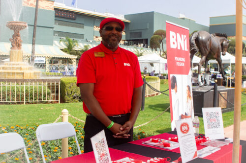 a man in a red BNI shirt and red hat standing beside a BNI pop up banner