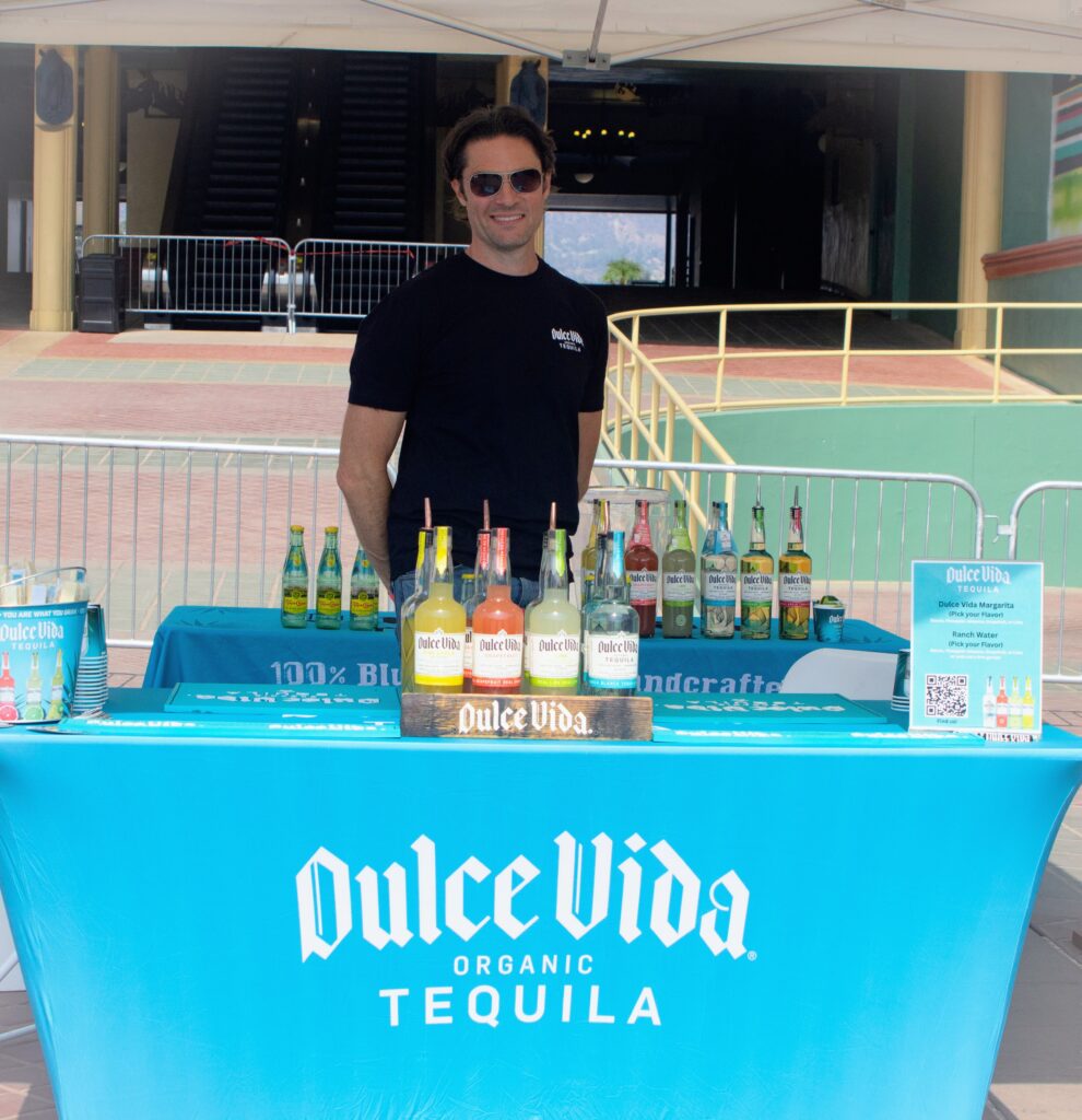 a man in a black tee shirt standing behind a booth for Dulce Vida Tequila set with a blue cloth with the logo