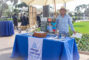 a woman in a blue shirt and straw hat standing behind a booth set with items for Always Best Care