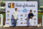 a woman in black and a hat standing to the right of a man in white, who is a good distance away from her, with the Taste of Arcadia Step and Repeat banner behind them