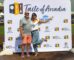 a woman in jeans and a white shirt with one leg and arm in the air standing with a man in a gray shirt and peach shorts in front of a step and repeat banner for Taste of Arcadia