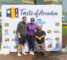 a woman in a purple shirt standing with two men in front of a step and repeat banner, one man is shorter than the other