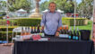 a man in a blue button up shirt standing behind a table set with bottles of wine