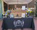 a bearded man standing behind a booth set with a black Mt Lowe table cloth and a cheers sign