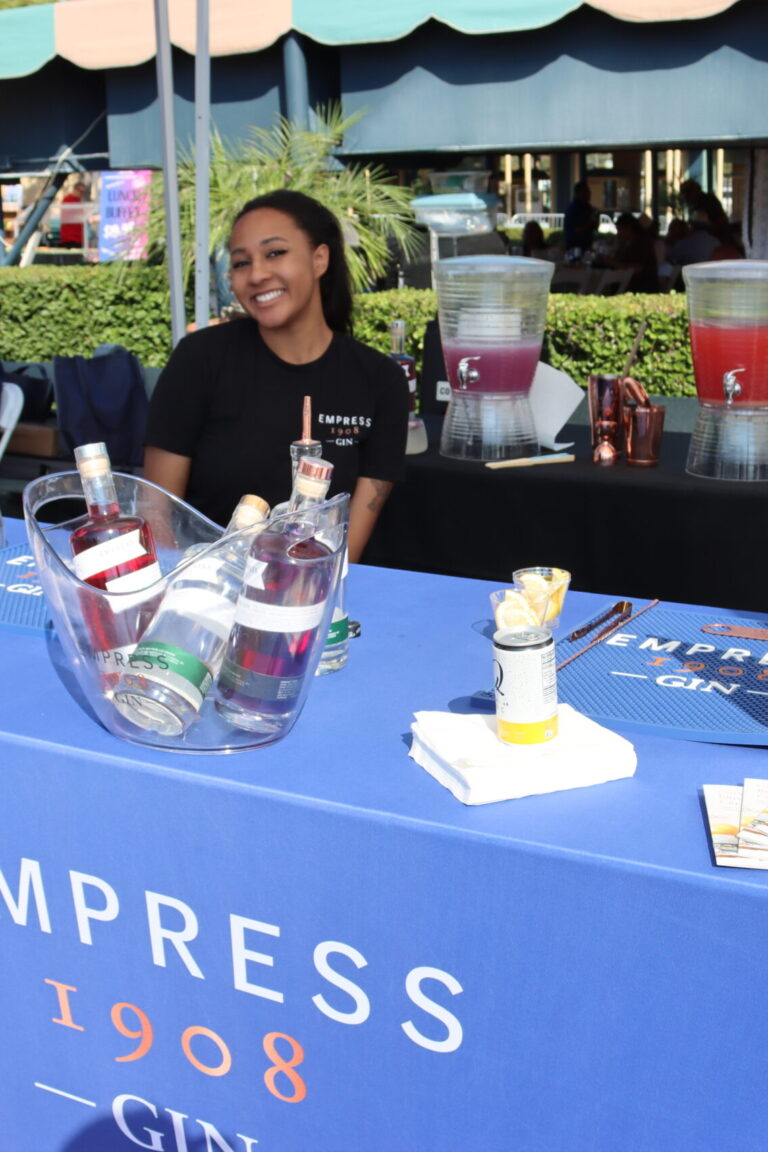a woman in a black shirt poses behind a table set with gin and a blue table cloth showing the Empress Gin logo