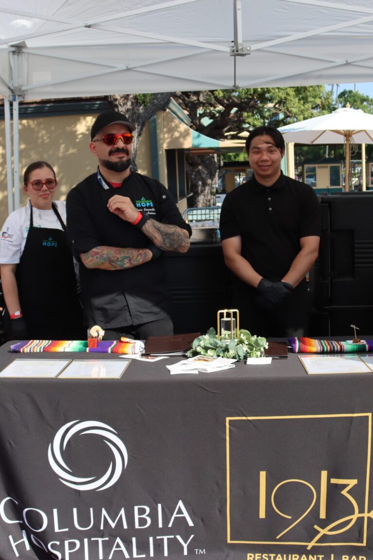 three men in black pose and give thumbs ups from behind a table set with a 1913 table cloth