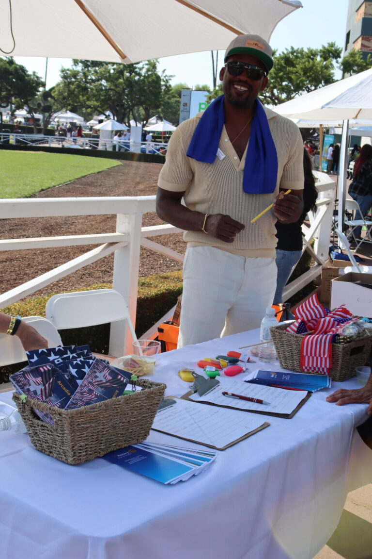 a woman in a yellow sundress poses beside a table set with items for Arcadia Rotary, with two men behind it