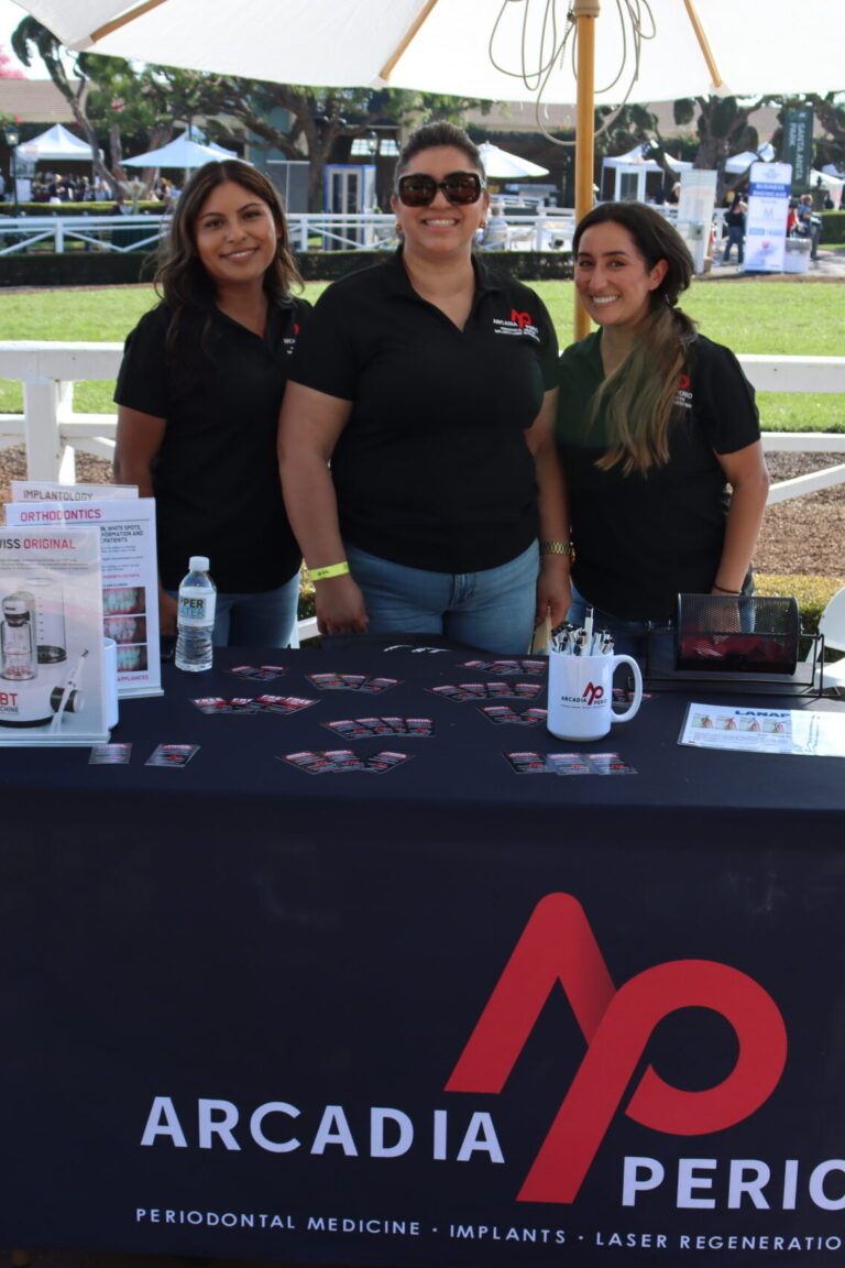 three women in black polos with the Arcadia Perio logo on them pose behind a booth set with an Arcadia Perio branded table cloth