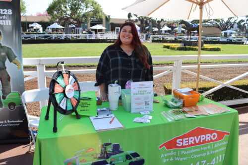 a woman with long, brown hair, in a plaid flannel, smiles behind a booth set with items for Servpro