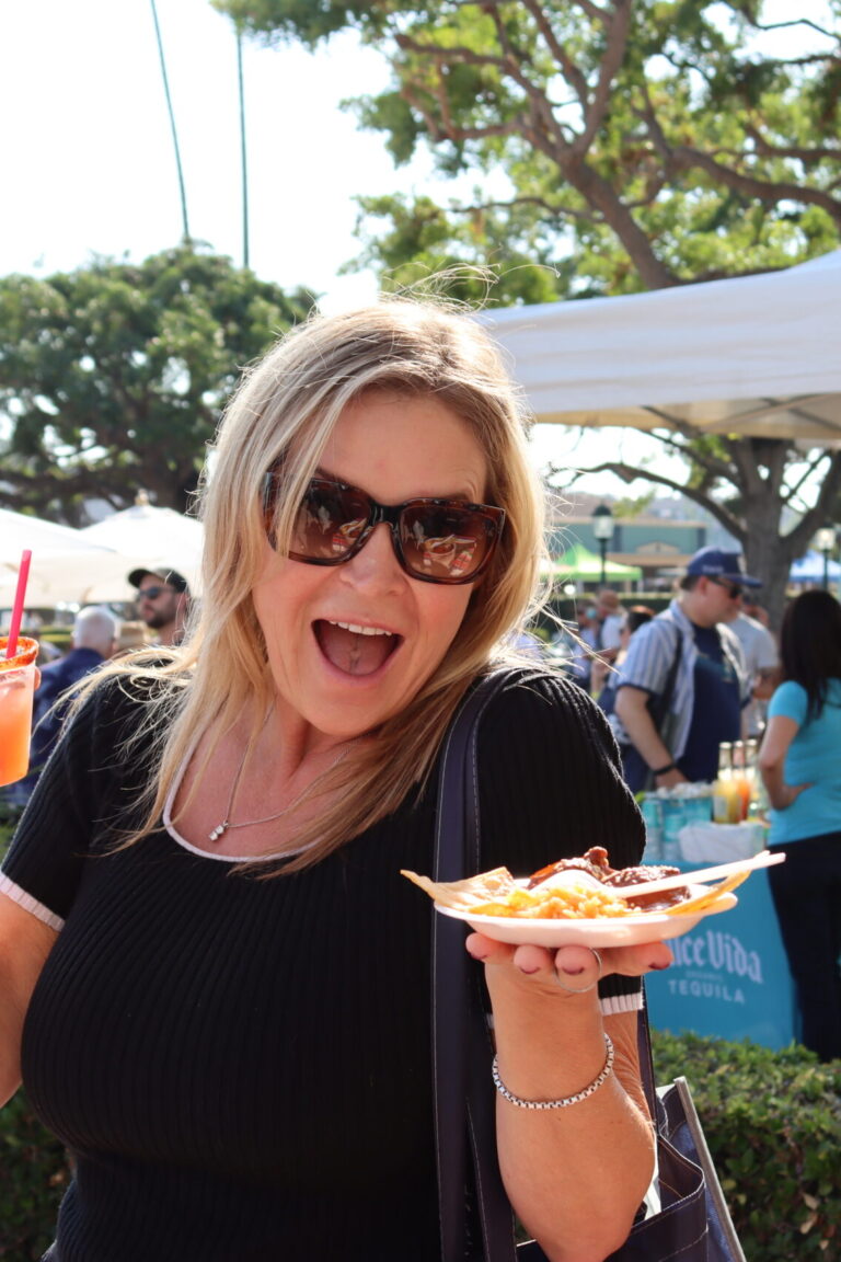a woman with long blonde hair, wearing a black t-shirt and sunglasses, holds up a cocktail and a plate of food while smiling broadly