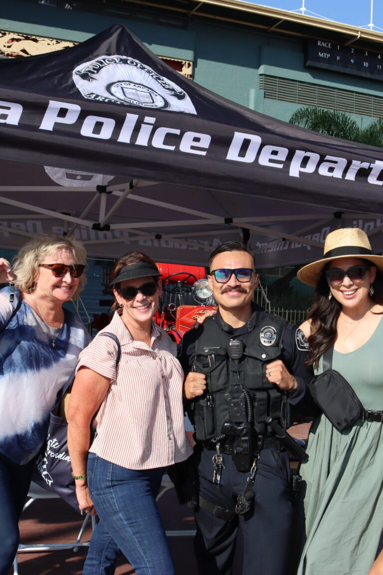 four women posing with a police officer under a tent that is labeled Arcadia Police Department