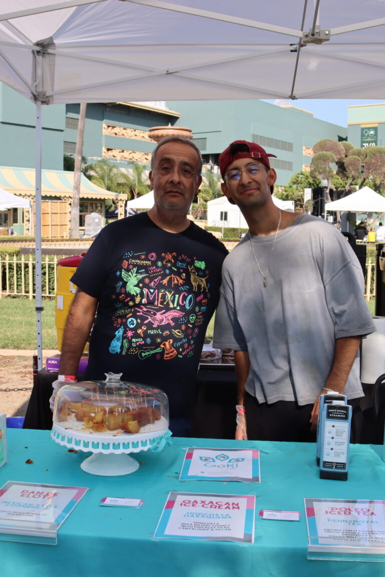 two men stand behind a table set with desserts under a glass dome and a turquoise blue table cloth
