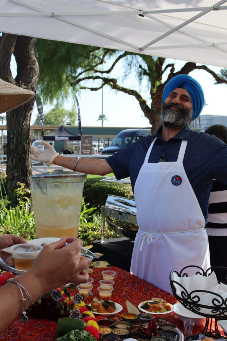 a man in blue shirt and white apron, wearing a blue turban, smile and spreads his arms wide while a woman in the foreground smiles and laughs