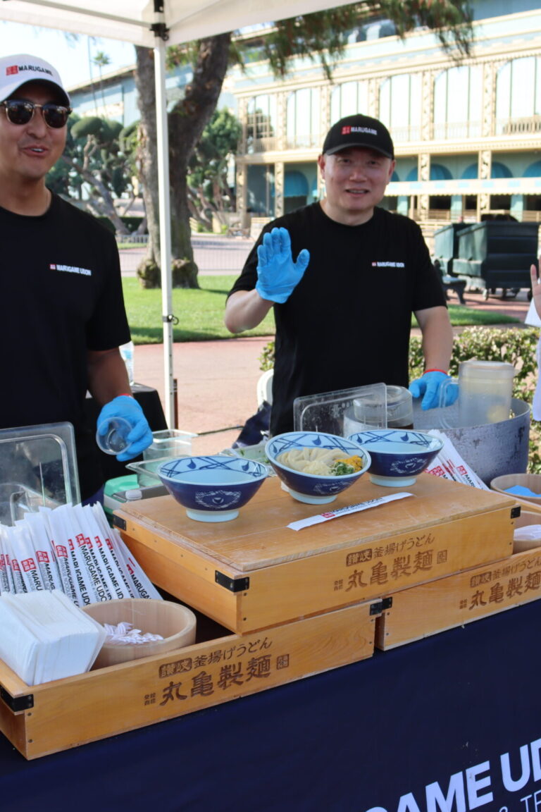 a woman and two men smile and wave behind a booth for Marugame Udon