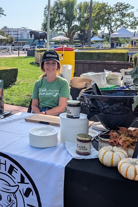 a woman in a black hat and green Twohey's shirt sits behind a booth set with fall items and a Twohey's overlay