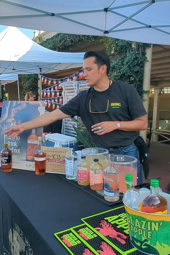 a man in a gray long sleeved shirt holding a Taste of Arcadia bag standing in front of a booth for Fireball, while a man in black points out a a bottle of Sazerac