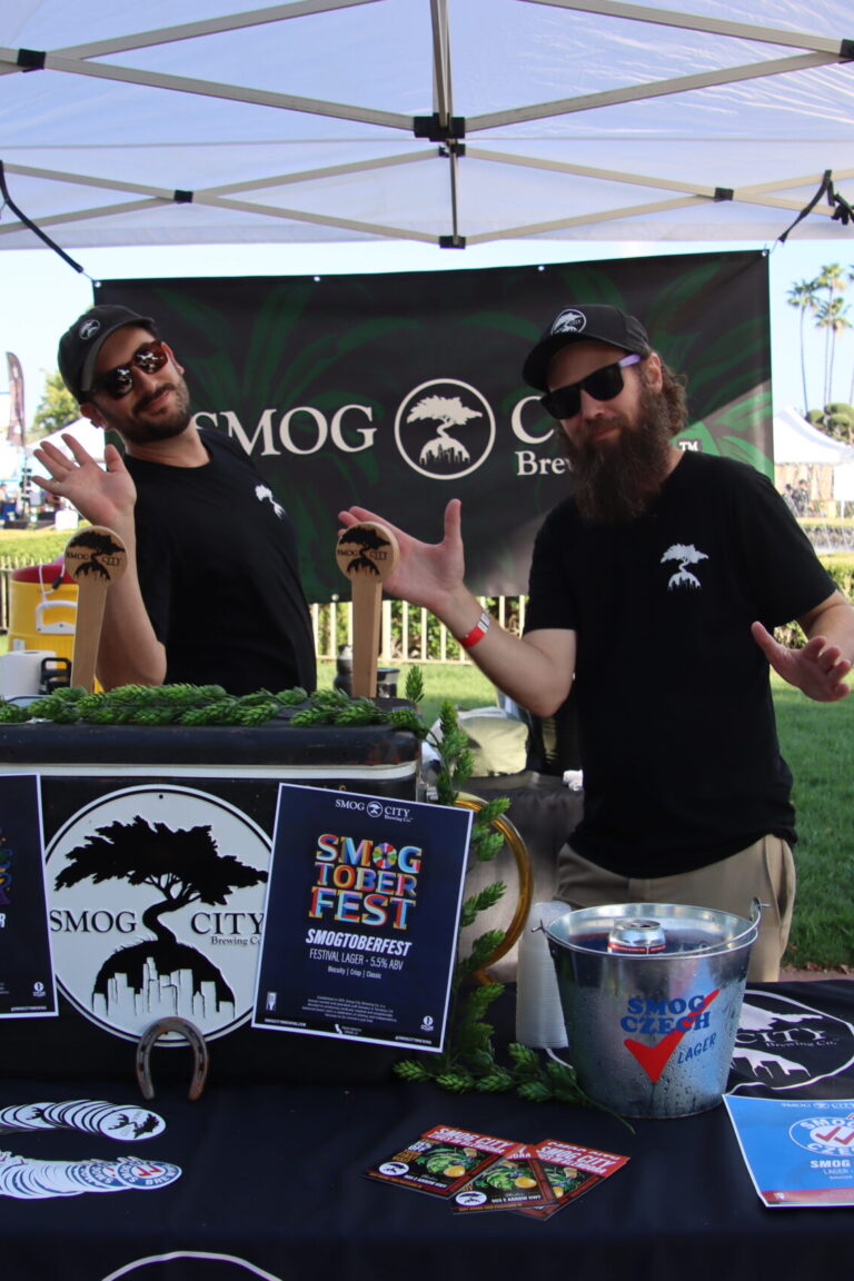 two men waving at the camera behind a booth for Smog City at Taste of Arcadia
