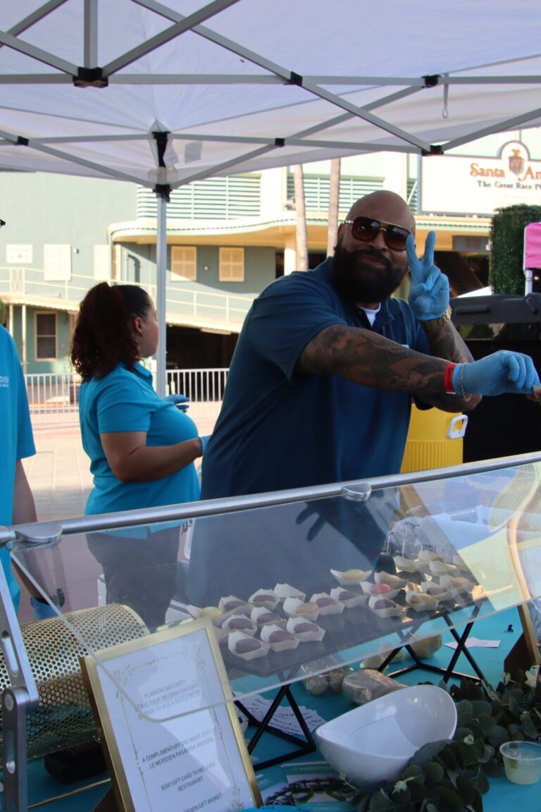 a man giving out a cookie and holding up a peace sign beside another man at the Le Meridien booth at Taste of Arcadia