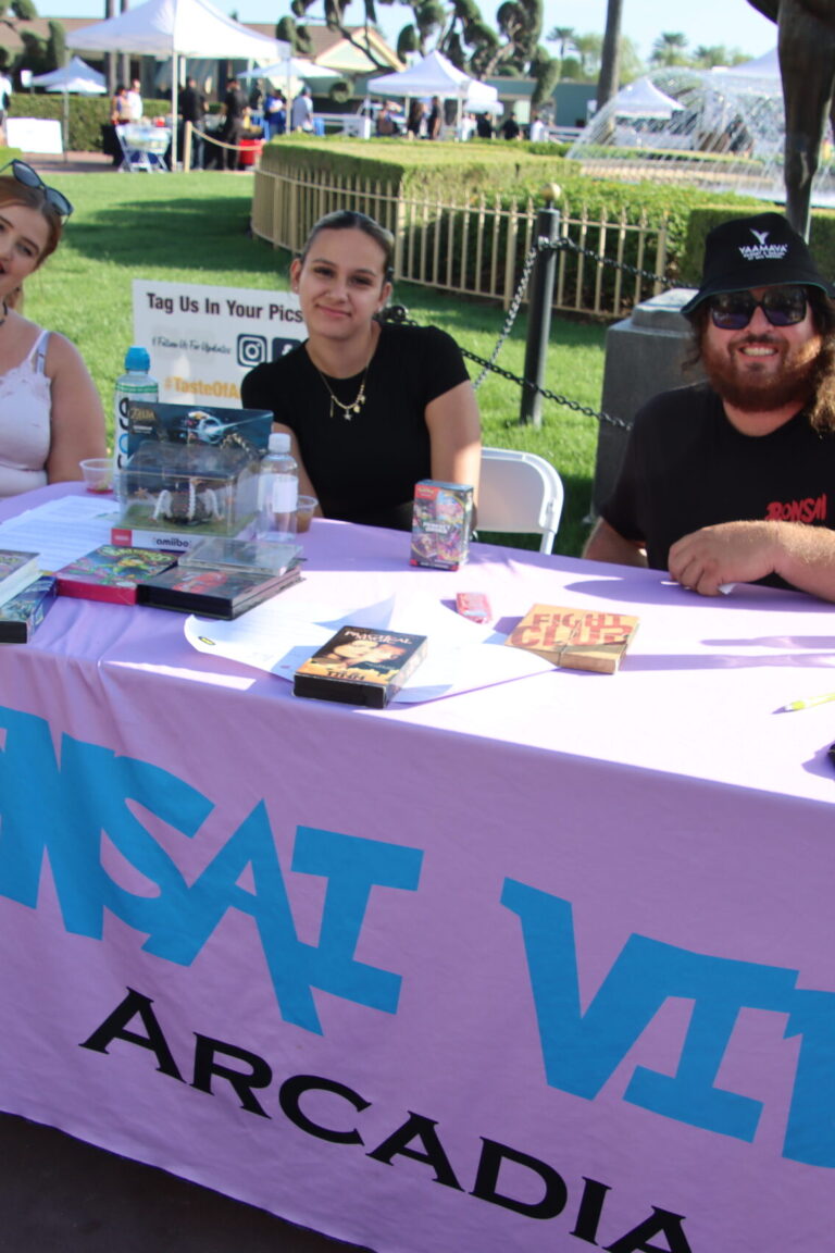 two women seated beside a man in a hat behind a booth for Bonsai Video at Taste of Arcadia