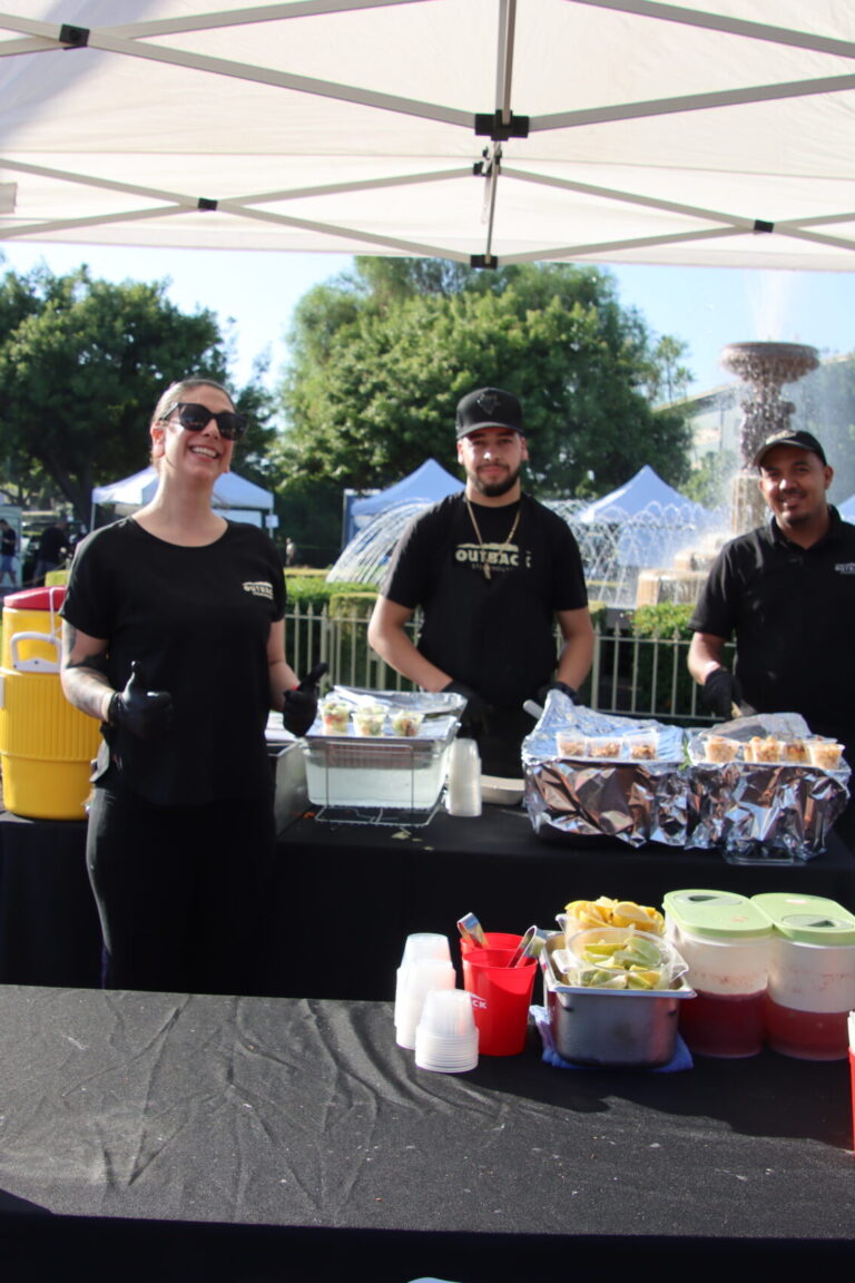 a woman giving thumbs up and standing in front of two men at a booth for Outback at Taste of Arcadia